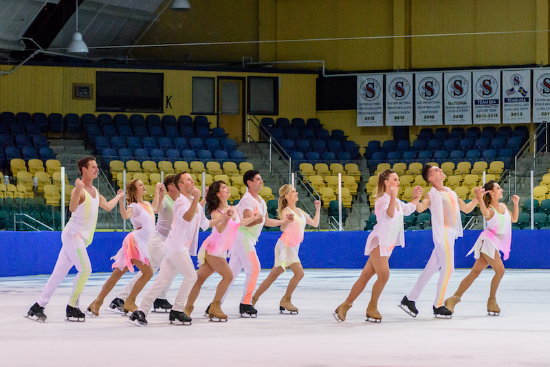 The entire IDI company in white costumes with pastel tie dye hold hands as if they're about to take a bow